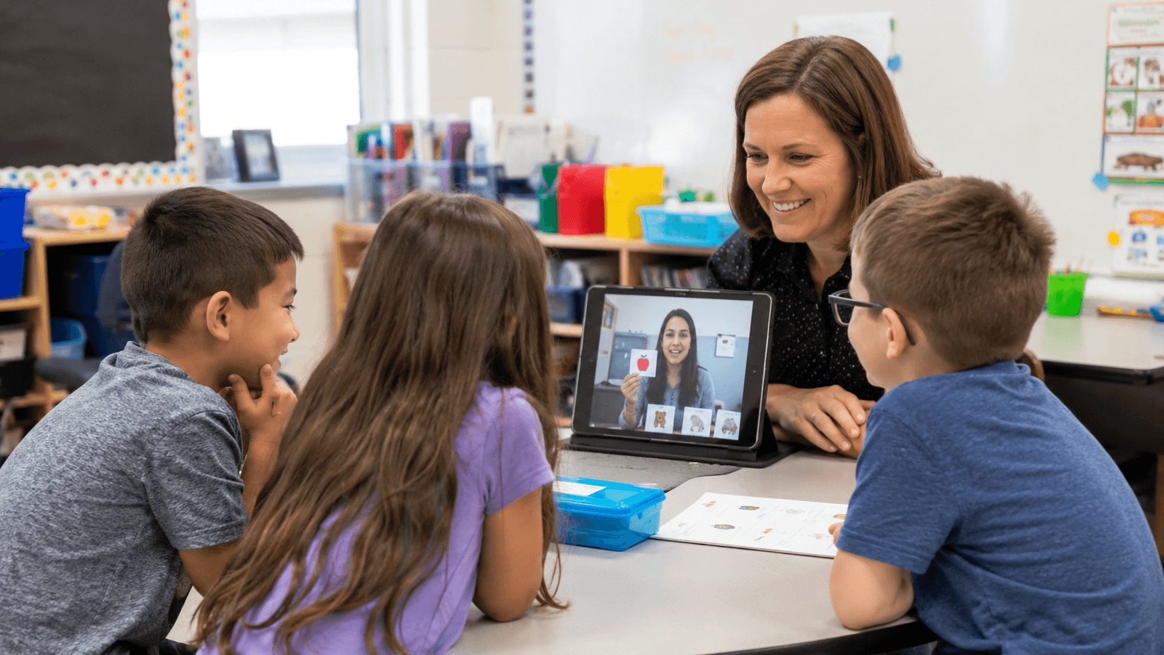 An educator and three students participate in a virtual learning or speech therapy session using a tablet in a classroom, while an online instructor displays educational picture cards during an interactive activity.